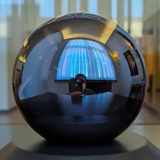 A highly reflective spherical object on display, with distorted reflections of a room and a person taking the photo.