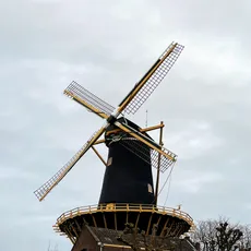 A traditional Dutch windmill with a black base, a band of decoration, and yellow blades against a cloudy sky in Woerden.