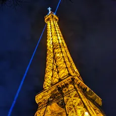 Illuminated Eiffel Tower at night, with a blue light beam from its peak against a dark blue sky, framed by faint tree branches.