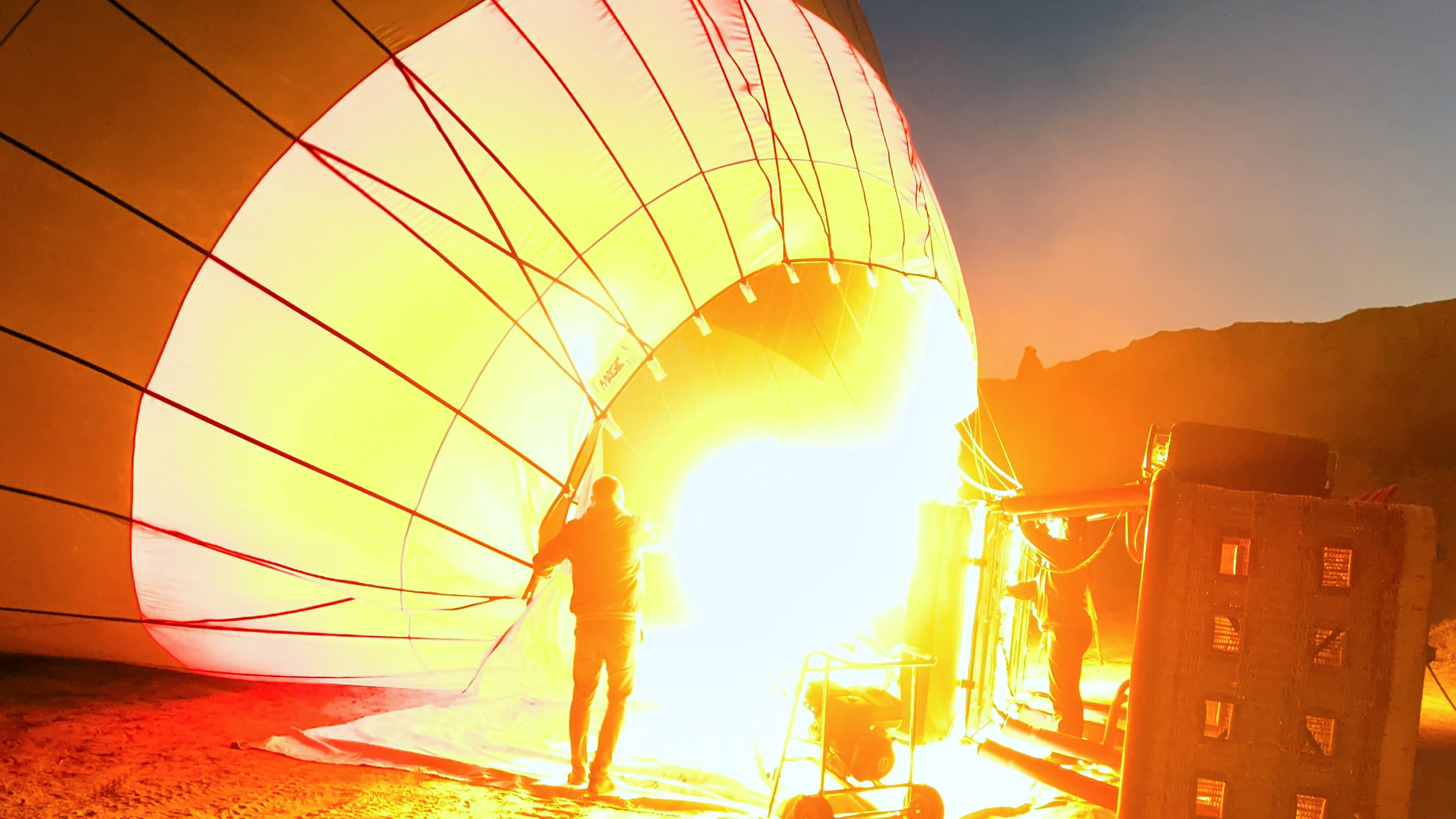 Silhouette of people by a hot air balloon being inflated, with a warm glow against a dusk sky.