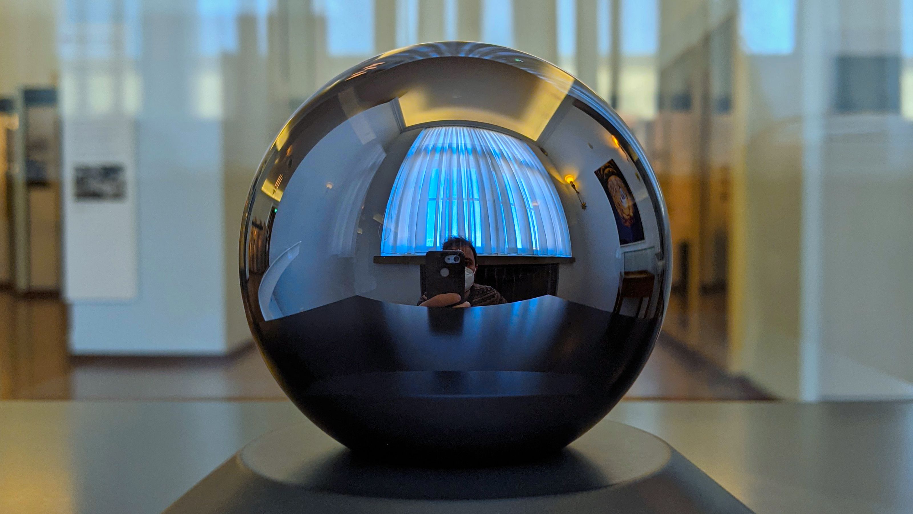 A highly reflective spherical object on display, with distorted reflections of a room and a person taking the photo.